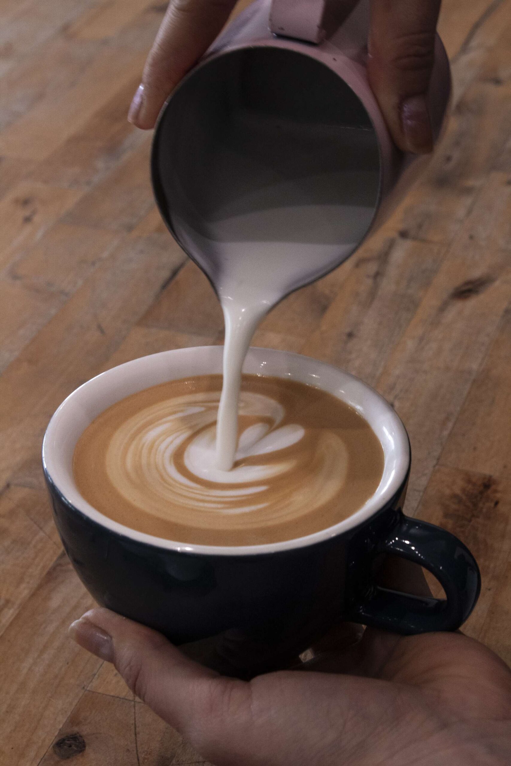 Barista pouring steamed milk into a cup of espresso to create latte art, held over a wooden counter