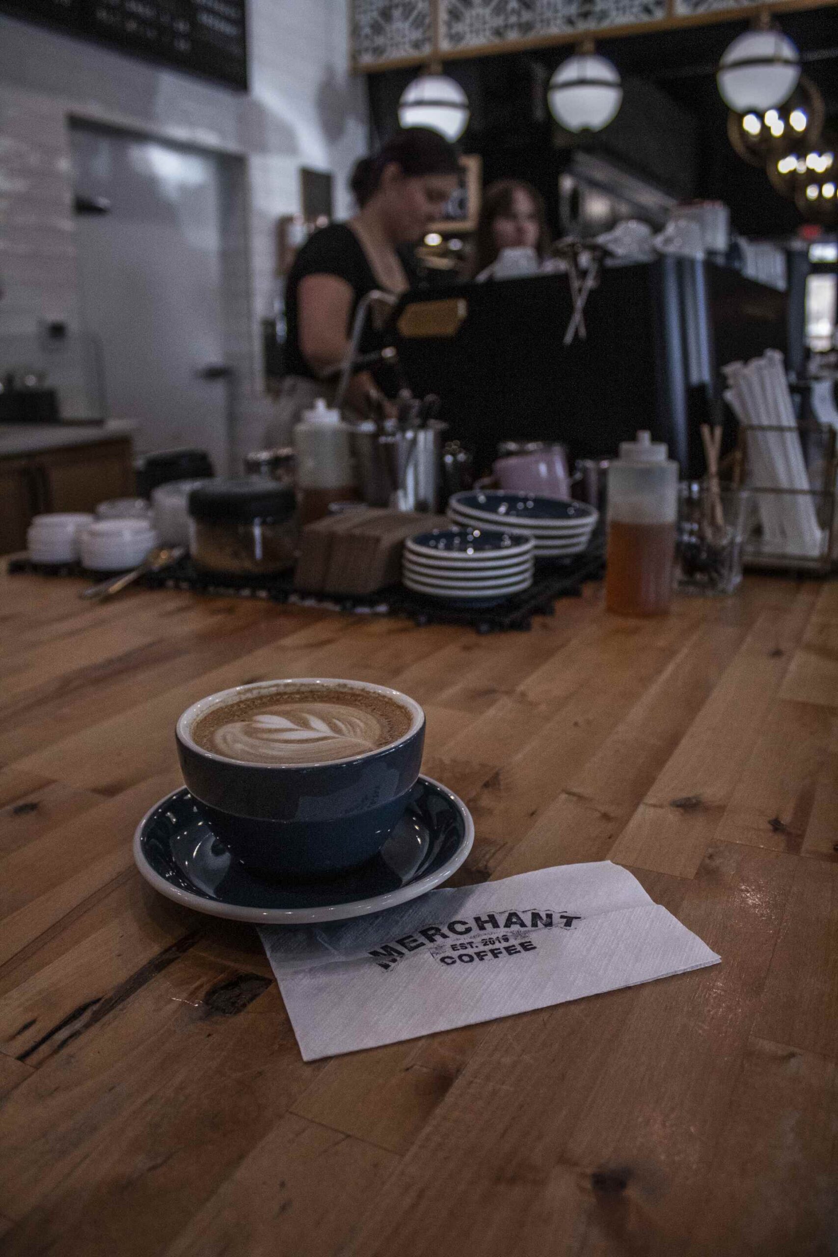 Latte with heart-shaped latte art on a wooden counter at Merchant Coffee, with baristas working behind the espresso machine.
