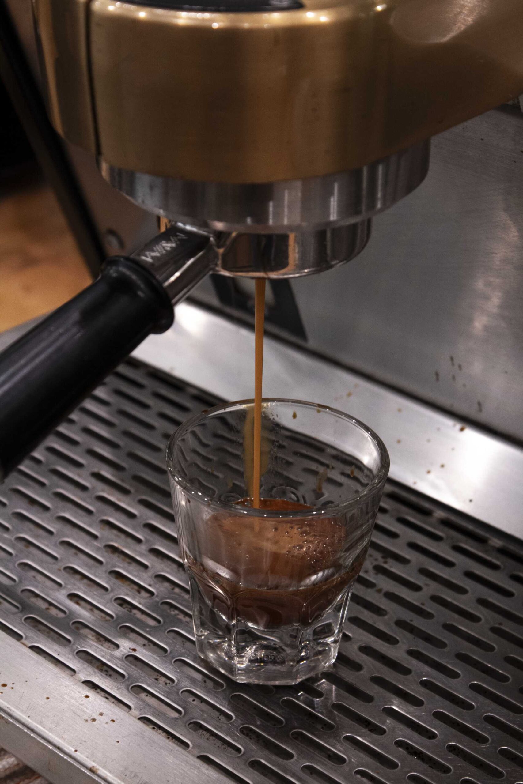 Espresso shot pouring from a commercial espresso machine into a clear glass shot glass on a metal drip tray.