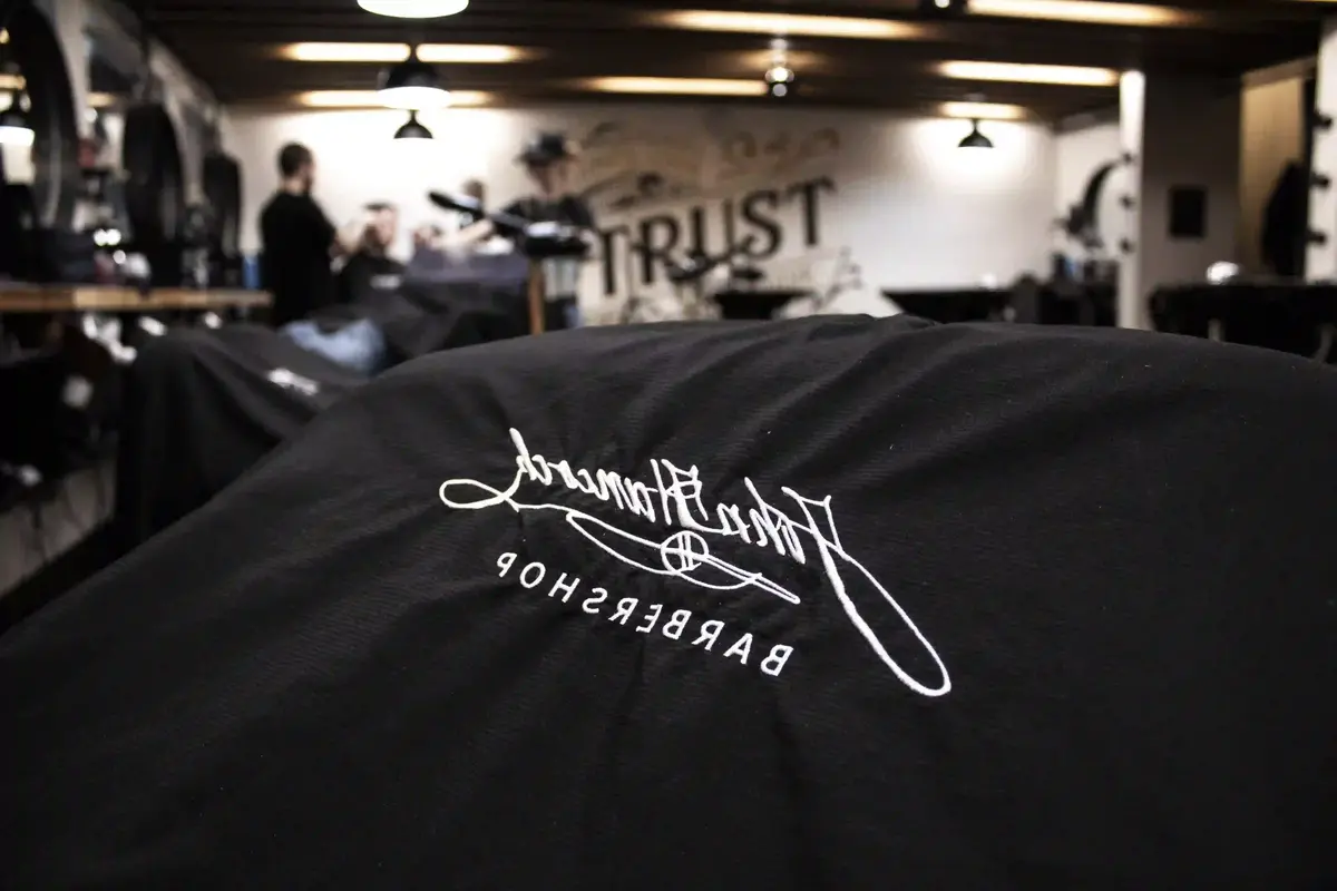 Haircut in progress at a barbershop Prescott location, with an embroidered barber cape in focus and the barber and client blurred in the background.