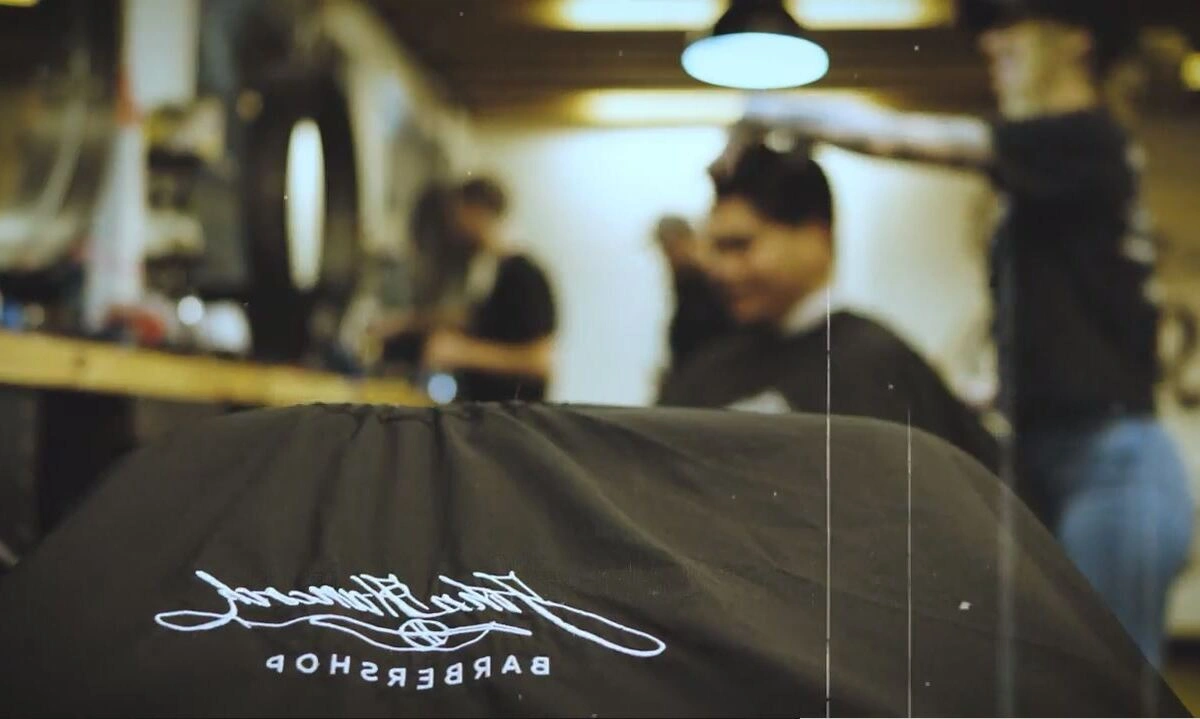 Haircut in progress at John Hancock barbershop Prescott location, with an embroidered barber cape in focus and the barber and client blurred in the background.