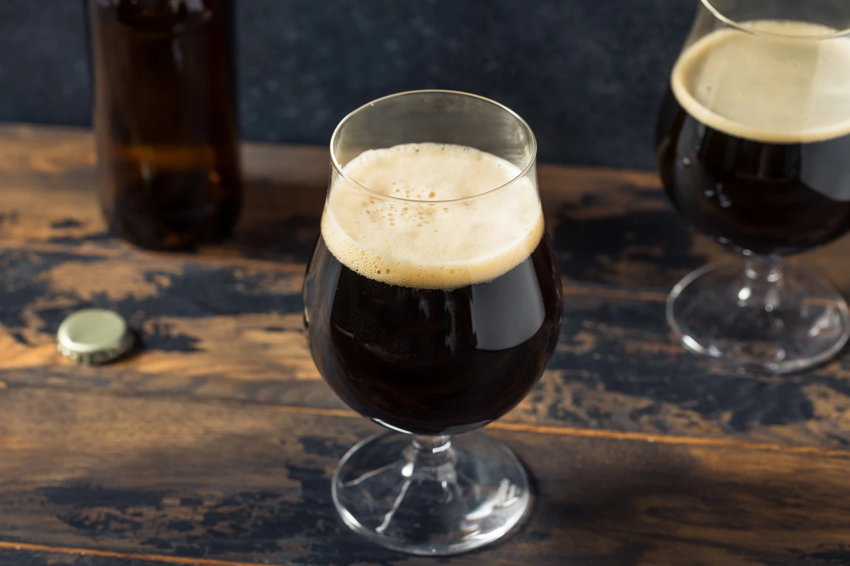 Dark Beers in a tulip glass with a creamy head on a wooden bar, with a bottle and cap in the background.